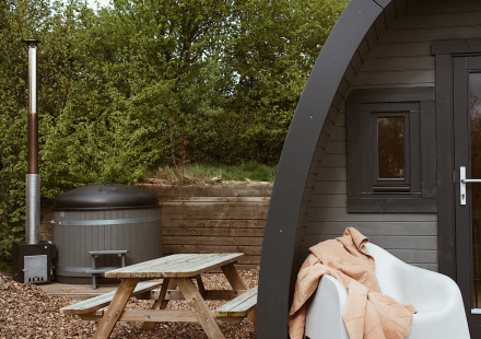 Glampingpod met hottub, picknicktafel en deken op stoel bij REBL Outdoor, Nederland, omgeven door natuur.