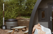 Glamping-Pod mit Whirlpool, Holztisch und Stuhl mit Decke bei REBL Outdoor, Niederlande, Naturkulisse.