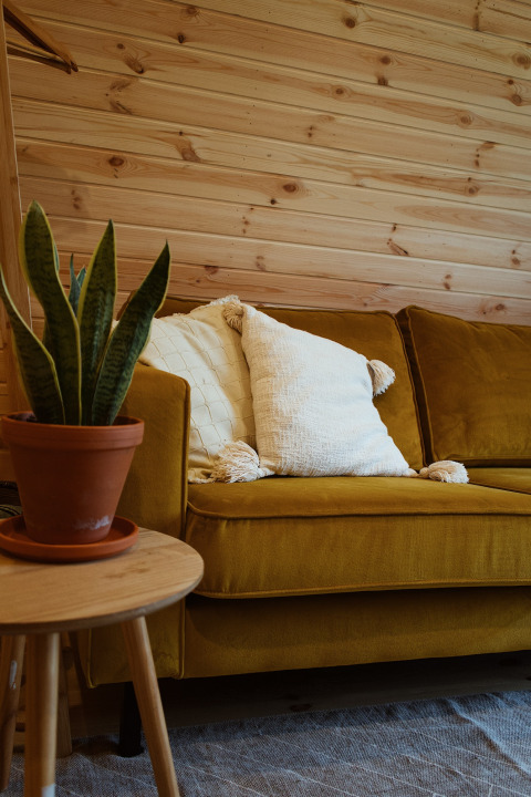 En hyggelig stue i en glamping pod med sennepsgul sofa, hvide puder, stuebirk og trævægge.
