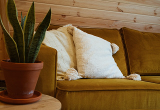 Knusse zithoek in een glampingpod met mosterdgele bank, witte kussens, plant en houten wand.
