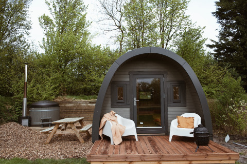 Glampingpod met gebogen dak, houten terras, stoelen, hottub en picknicktafel in het groen.