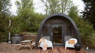 Glampingpod met gebogen dak, houten terras, stoelen, hottub en picknicktafel in het groen.