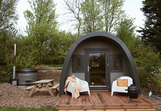 Glampingpod met gebogen dak, houten terras, stoelen, hottub en picknicktafel in het groen.