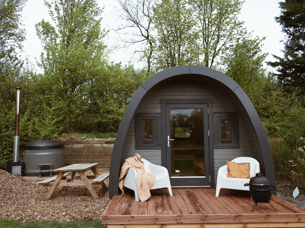 Glamping pod met gebogen dak, houten terras, stoelen, hottub en picknicktafel in groene omgeving.