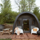 Glamping pod med buet tag, træterrasse, stole, boblebad og picnicbord omgivet af grønne træer.