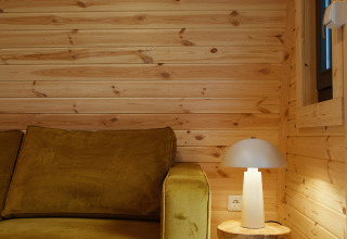 Binnenzicht van glamping pod bij REBL Outdoor Nederland met gele sofa, houten tafeltje en lamp.