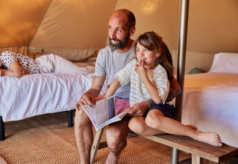 En far og datter sidder sammen på en bænk i en glamping-tipi, læser en bog, afslappet stemning.