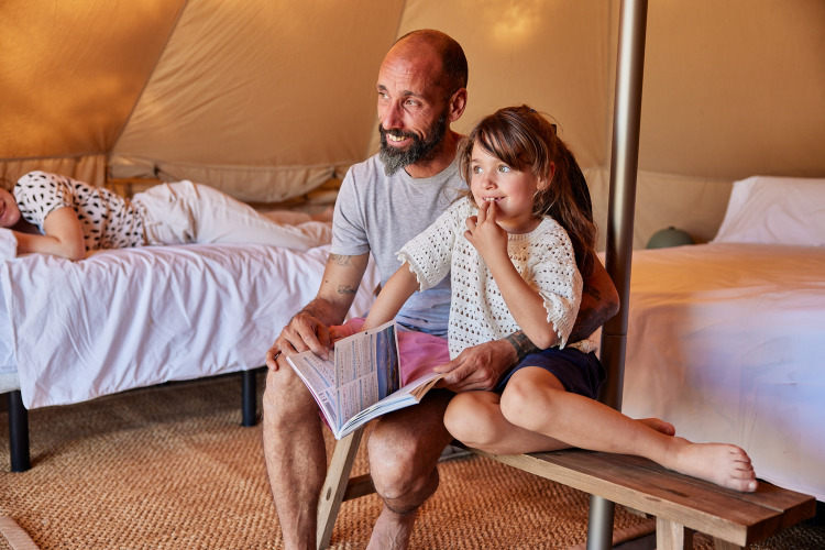 Padre e hija sentados en un banco leyendo un libro dentro de una tienda Glamping Bell Tent en España.