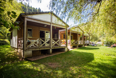Chalet confort (river view) wooden cabins by the river, nestled in lush green surroundings and sunlight.