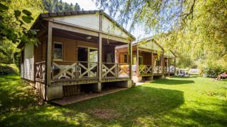 Chalet confort (river view) wooden cabins by the river, nestled in lush green surroundings and sunlight.