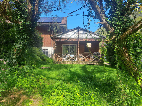 Chalet confort con terrazza in legno, verde rigoglioso, sole, cielo azzurro e vista sul ruscello.