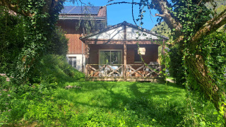 Chalet confort with wooden terrace, lush greenery, sunshine, blue sky and stream view from the garden.