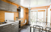 Moderne chaletkøkken og spisestue med trævægge og store vinduer, udsigt til terrasse og natur ved en bæk.