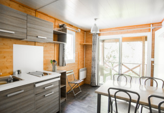 Moderne chaletkøkken og spisestue med trævægge og store vinduer, udsigt til terrasse og natur ved en bæk.