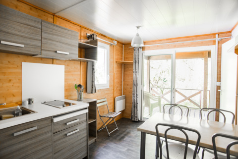 Moderne chaletkeuken en eetruimte met grote ramen, terras en uitzicht op het beekje buiten.