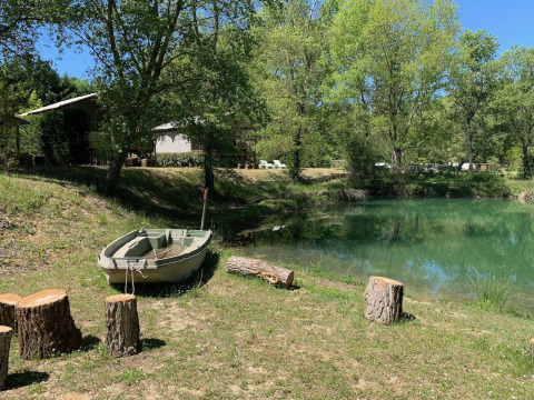 Barque au bord d’une mare paisible, troncs pour s’asseoir et arbres au Flower Camping La Rivière en France.