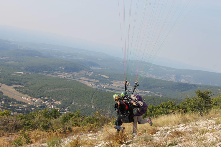 Two people tandem paragliding from a hillside with scenic views near Flower Camping La Rivière in France.