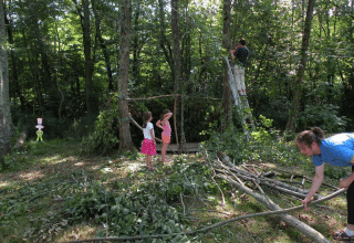 Famiglia costruisce un rifugio con rami nel bosco al Camping Le Périgord, Francia, adulti e bambini collaborano.