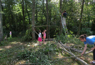 Familie bygger hytte med grene i en skov på Camping Le Périgord, Frankrig, børn og voksne samarbejder.