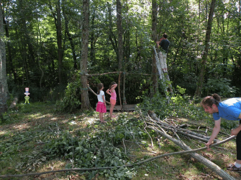 Famiglia costruisce un rifugio con rami nel bosco al Camping Le Périgord, Francia, adulti e bambini collaborano.