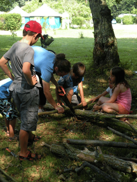 Children and adult sawing branches together in the shade at Camping Le Périgord, Nouvelle-Aquitaine, France.