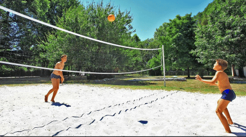 Due bambini giocano a pallavolo su un campo di sabbia circondato da alberi a Camping Le Périgord, Nouvelle-Aquitaine, Francia.