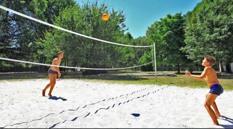 Dos niños juegan al voleibol en una cancha de arena rodeada de árboles en Camping Le Périgord, Nouvelle-Aquitaine, Francia.