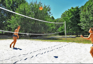 Two kids playing volleyball on a sandy court surrounded by green trees at Camping Le Périgord, Nouvelle-Aquitaine, France.