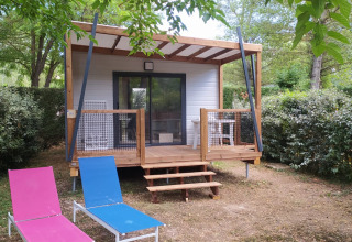 Chalet Cajou Confort met houten veranda en loungestoelen in de tuin, verscholen tussen groene struiken.