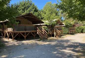 Glamping tenten op verhoogde houten vlonders tussen bomen en groen, onder een heldere blauwe lucht.