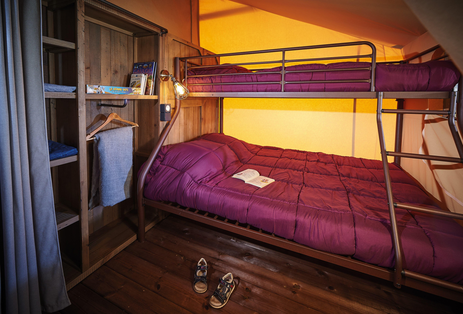 Knusse glampingkamer met stapelbed, paarse dekbedden, houten kast en een open boek op het bed.