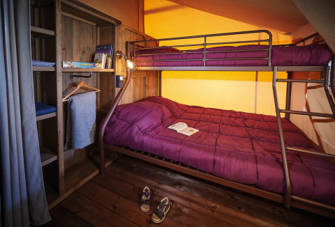 Habitación de glamping con litera, ropa de cama morada, estantería de madera y libro abierto.