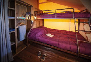 Gezellige glampingkamer met een stapelbed, paarse dekens, houten kast en een open boek op bed.