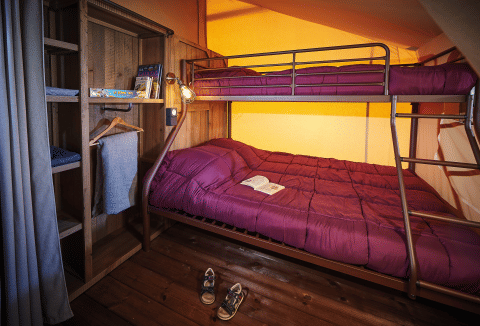 Camera glamping accogliente con letto a castello, piumone viola, armadio in legno e libro aperto.