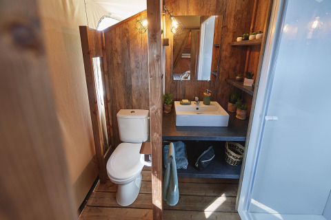 Baño dentro de una tienda safari con paneles de madera, inodoro, lavabo y estanterías en Flower Camping La Rivière.