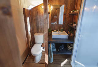 Baño dentro de una tienda safari con paneles de madera, inodoro, lavabo y estanterías en Flower Camping La Rivière.