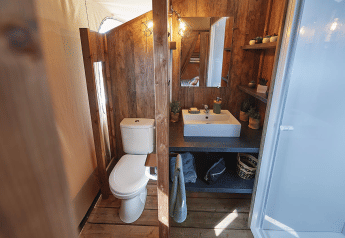 Bathroom inside a safari tent featuring wood paneling, toilet, sink, mirror and shelves at Flower Camping La Rivière.