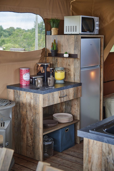 Pequeña cocina acogedora en tienda safari con acabado de madera, microondas, nevera y vajilla en Francia.