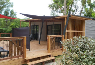 Afbeelding van een glampingverblijf met houten terras, zonnezeilen en moderne buitenzit ruimte in het groen.