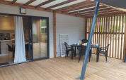 Porche cubierto de madera con mesa y sillas de comedor frente a un alojamiento glamping con cocina abierta.