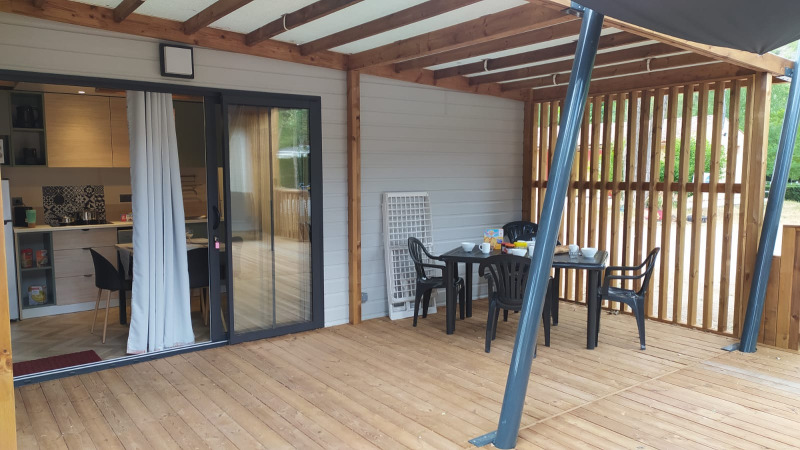 Porche cubierto de madera con mesa y sillas de comedor frente a un alojamiento glamping con cocina abierta.