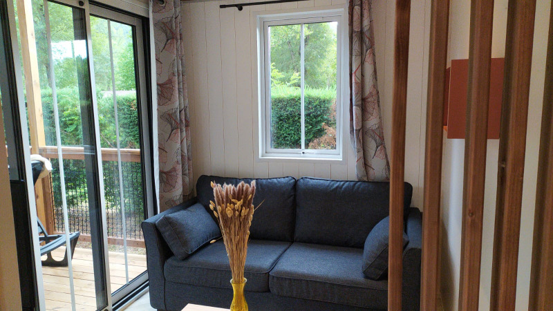Petit salon lumineux avec canapé sombre, fenêtre, rideaux et vue sur le jardin, dans un logement glamping.