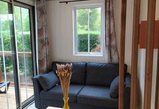 Gezellige zithoek met donkere sofa, raam, gordijnen en tuinzicht in een glamping-accommodatie.