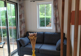 Acogedora sala con sofá oscuro, ventana, cortinas de hojas y vista al jardín en alojamiento glamping.