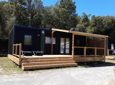 Zwarte glampinghut met groot houten terras en overdekte zone, gelegen in een bosrijke, natuurlijke omgeving.