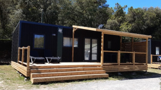 Schwarze Glamping-Unterkunft mit großer Holzterrasse und Dach, umgeben von Bäumen und Natur.