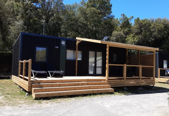 Cabaña negra de glamping con amplia terraza de madera y zona cubierta, rodeada de árboles y naturaleza.