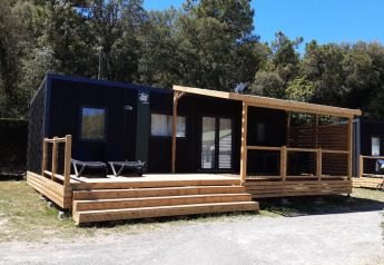 Cabane de glamping noire avec grande terrasse en bois et espace couvert, entourée de forêt et de nature.