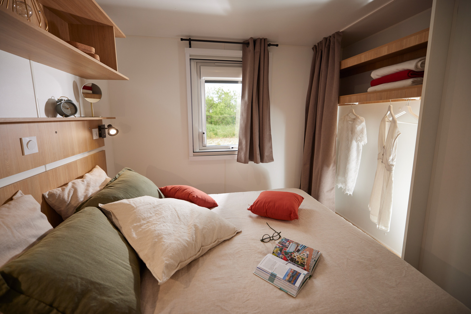 Knusse glamping slaapkamer met tweepersoonsbed, kussens, open kledingkast en raam met uitzicht.