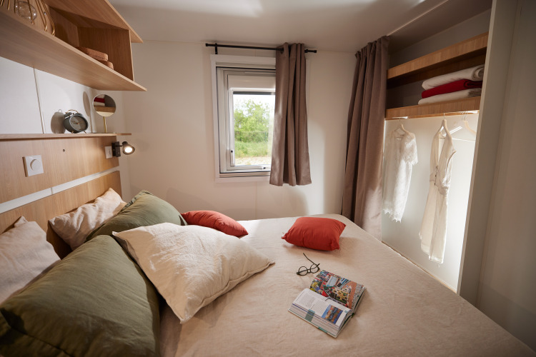 Modernes Glamping-Schlafzimmer mit Doppelbett, Kissen, Buch, Fenster und offenem Kleiderschrank.